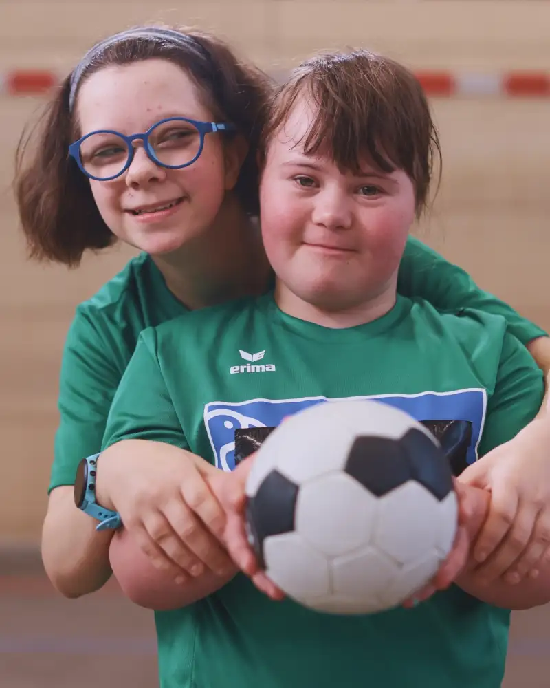 Zwei Kinder stehen in grünen Trikots in einer Sporthalle. Das eine Mädchen steht hinter dem anderen. Beide halten zusammen einen Ball fest. Im Hintergrund sieht man unscharf ein Handball-Tor.