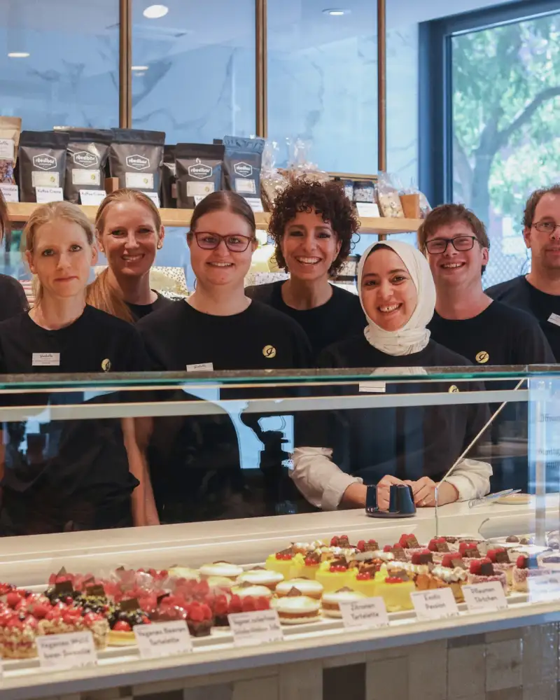Eine Gruppe von Mitarbeitenden einer Konditorei steht hinter der Theke und lacht in die Kamera. Die Personen sind unterschiedlicher ethnischer Herkunft und mit und ohne sichtbare Behinderung.