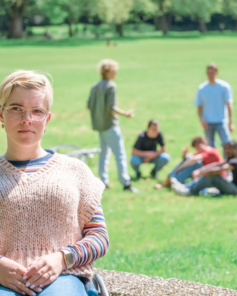 Eine junge Frau im Rollstuhl schaut in die Kamera. Im Hintergrund sieht man auf einer Wiese mehrere Jungedliche.