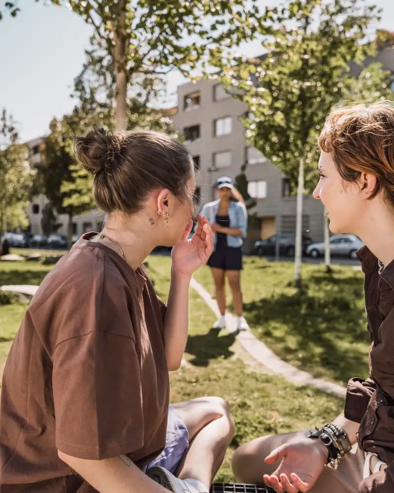 Zwei Jugendliche sitzen im Park und schauen von der Kamera weg. Sie schauen auf eine Person, die in Richtung Kamera auf einem Weg entlang geht.