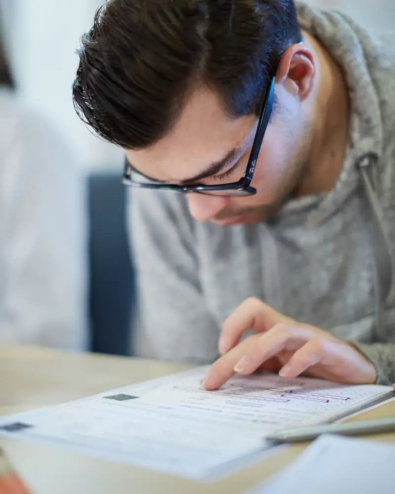 Ein junger Mann mit Brille beugt sich über ein Papier, das auf einem Tisch liegt. Seinen Finger nutzt er als Lesehilfe.