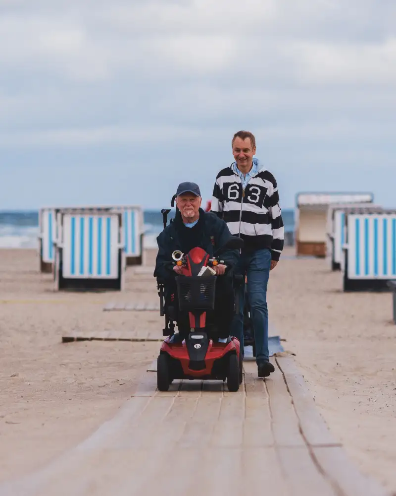 Zwei Männer gehen an einem Strand entlang. Einer der beiden nutzt einen Rollstuhl. Im Hintergrund sieht man das Meer sowie Strandkörbe.