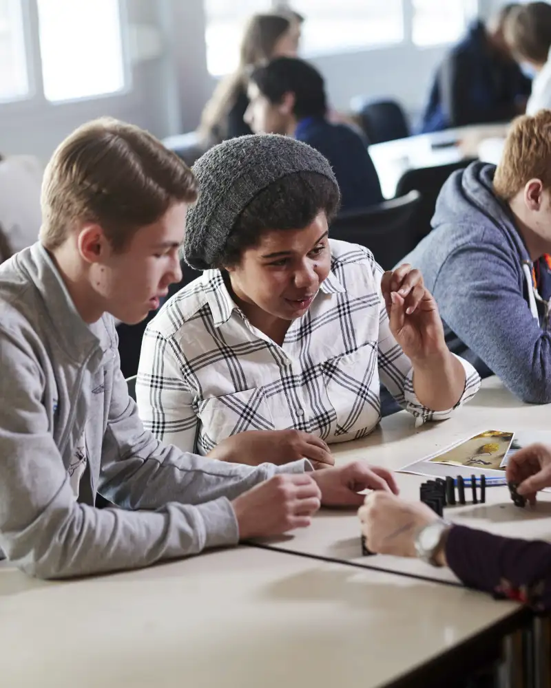 Jugendliche sitzen an langen Tischreihen. Fokus des Bilds sind zwei von ihnen. Die übrigen sieht man im Hintergrund.