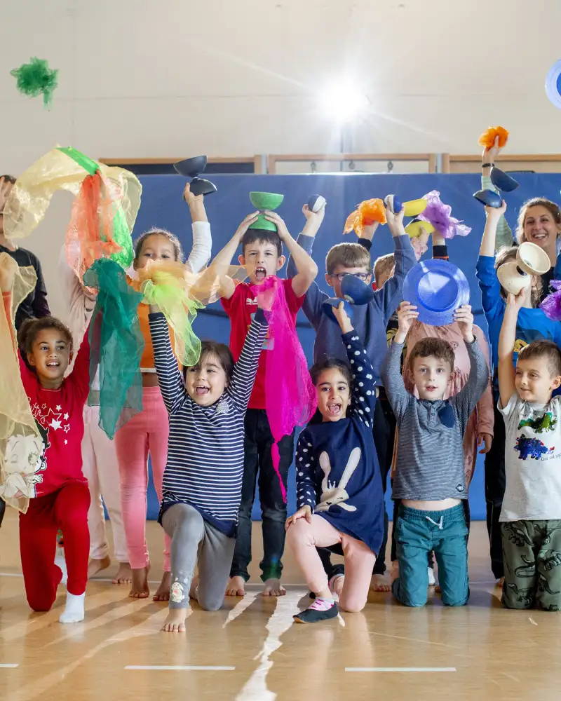 Eine Gruppe von Kindern in einer Turnhalle. Alle jubeln und halten bunte Tücher, Diabolos und Bälle in die Luft.