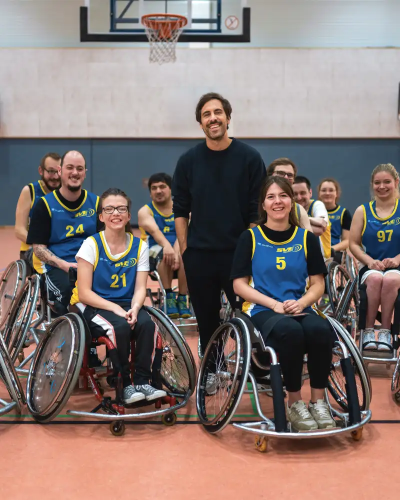 Max Giesinger steht in einer Turnhalle in mitten einer Gruppe Rollstuhl-Basketballer*innen