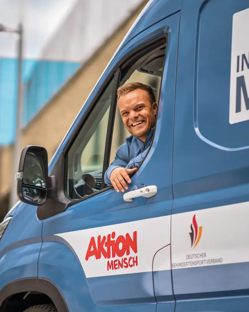 In einem blauen Kleintransporter sitzt ein kleinwüchsiger, sportlicher Mann mit kurzen blonden Haaren. Er sieht aus dem Fenster der Fahrertür und lacht in die Kamera. Auf dem Bus sind Ausfkleber angebracht. Auf einem steht "Inklusionsmobil". Auf den anderen sind die Logos der kooperatiosnpartner zu sehen, die diesen Bus unterstützen: Aktion Mensch, Deutscher Behindertensportverband und REWE.