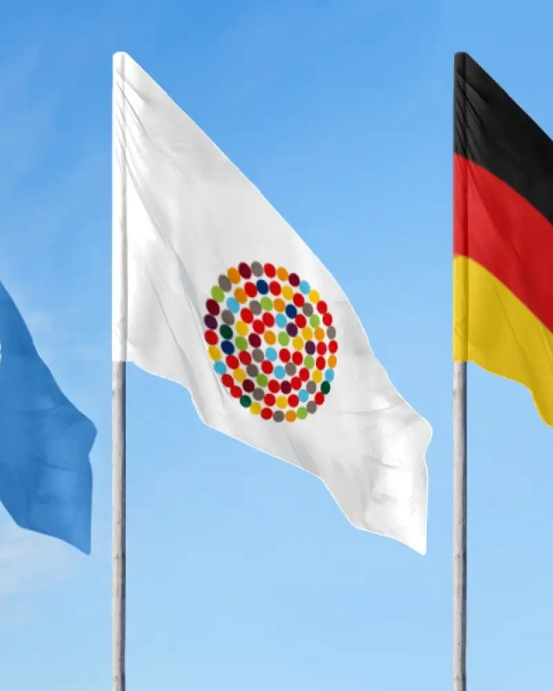 Drei Flaggen an Mast vor blauem Himmel: Eine mit dem Logo der UN, eine mit vielen bunten Punkten im Kreis, die Inklusion symbolisieren sollen und die deutsche Fahne