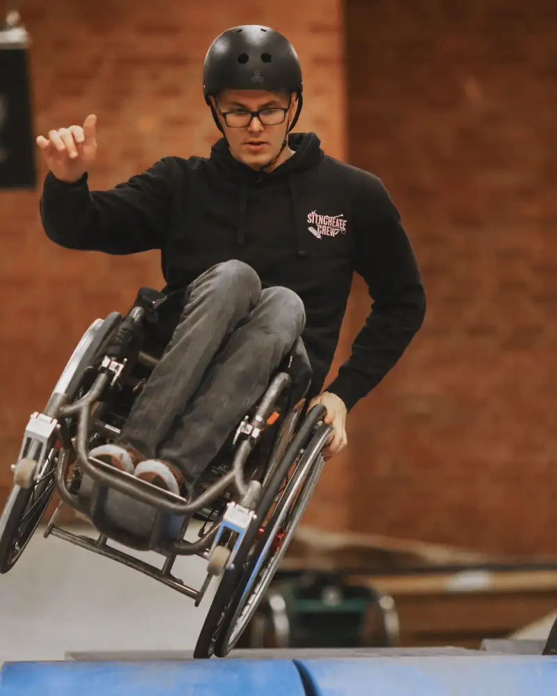 Ein Mann im Rollstuhl fährt mit einem Rollstuhlrad in einer Turnhalle über eine Matte. Das andere Rollstuhlrad berührt nicht den Boden.