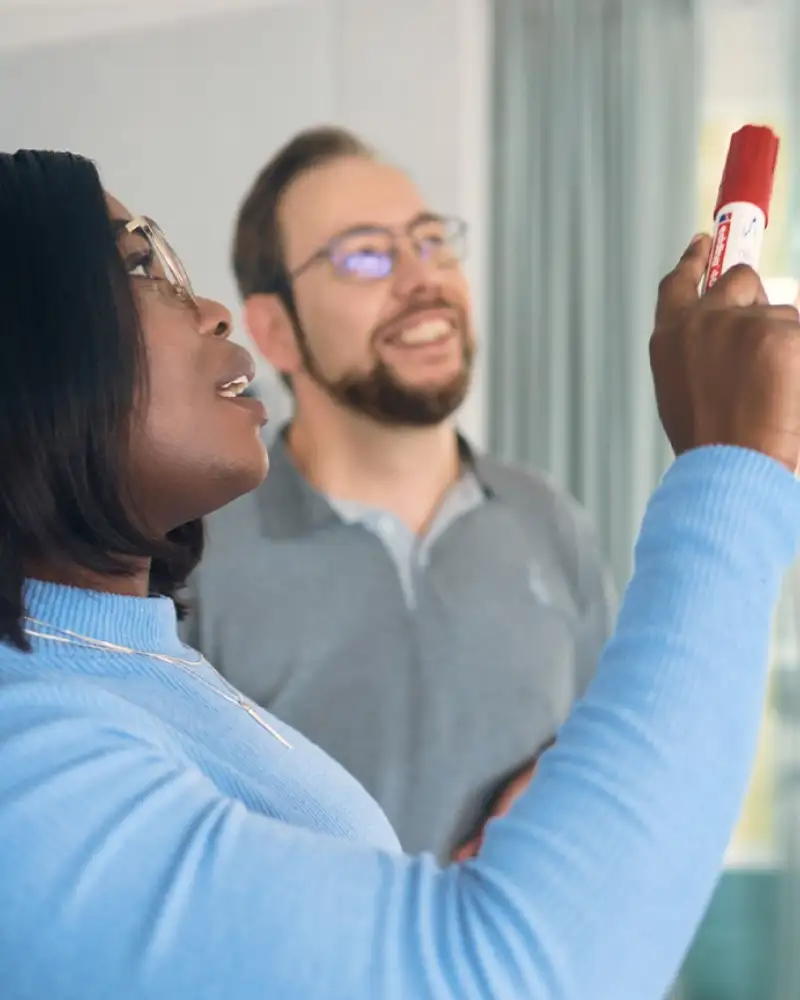 Eine Frau und ein Mann stehen gemeinsam vor einer Gläsernen Wand. Die Frau hält einen Stift in der Hand und zeigt damit auf einen pinken Klebezettel, der vor ihr an der Glasscheibe klebt.