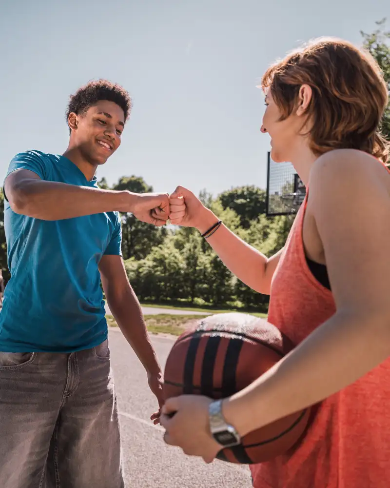 Zwei Jugendliche stehen auf einem Basketballplatz gegenüber und begrüßen sich freundschaftlich mit dem Faustgruß.