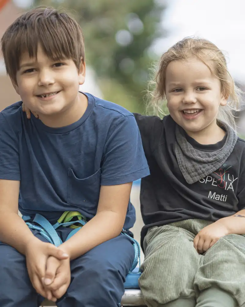 Zwei Jungs sitzen nebeneinander auf dem Rand eines Trampolins. Der kleinere hat seinen Arm freundschaftlich auf die Schulter des anderen gelegt. Beide tragen T-Shirts lächeln fröhlich in die Kamera.