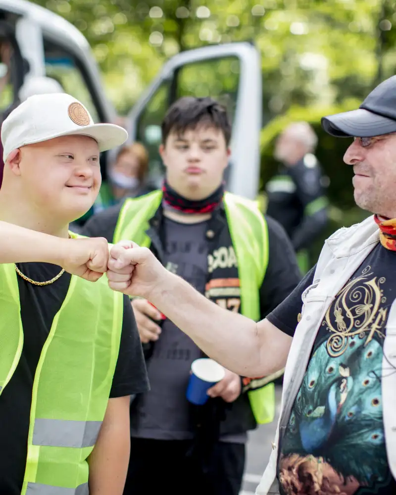 Zwei Biker begrüßen sich mit einem freundschaftlichen Faustgruß. Einer von ihnen hat das Down-Syndrom und trägt eine Warnweste. Der andere trägt ein schwarzes Basecap und ein buntes Halstuch.