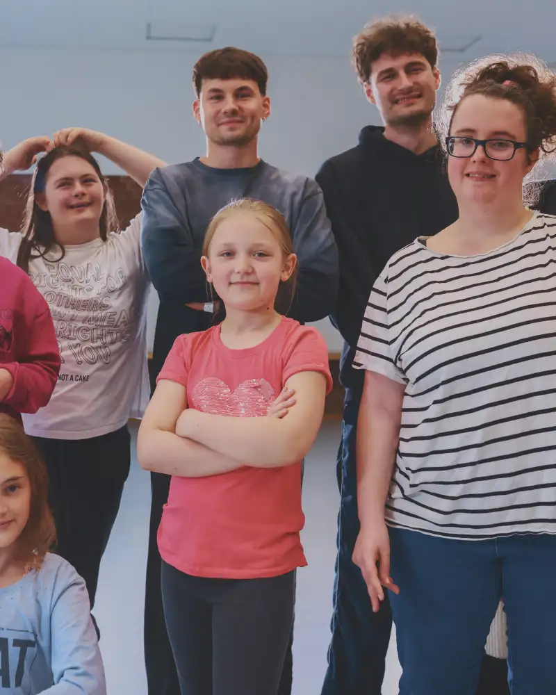 Eine achtköpfige Gruppe von Jungen und Mädchen, in der Altersspanne von etwa 10 bis ca. 25 posieren zusammen in einem Tanzstudio für das Foto.