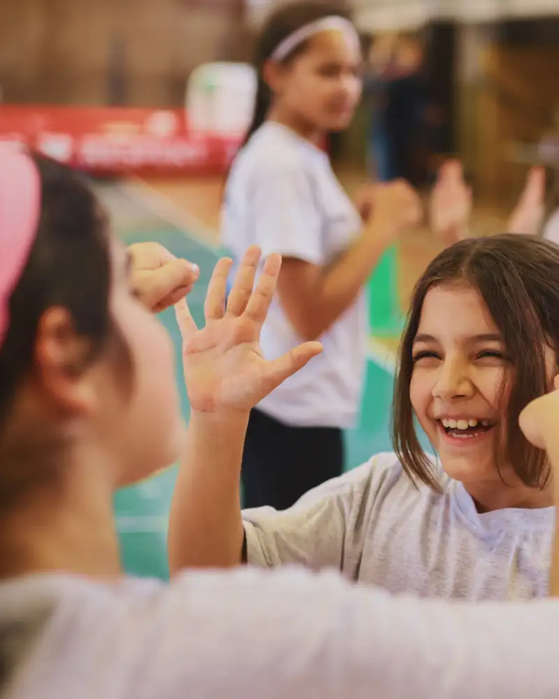 In einer Turnhalle klatschen sich zwei Mädchen ab. Im Hintergrund sind unscharf zwei weitere Kinder zu sehen. Auch sie klatschen sich ab.