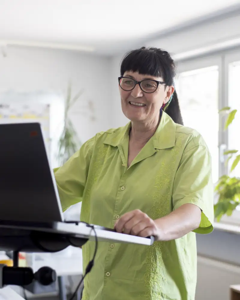Eine Frau steht in einem Büroraum an einem Laptop. Sie trägt einen Brille, einen Pferdeschwanz und eine hellgrüne, kurzärmelige Bluse.