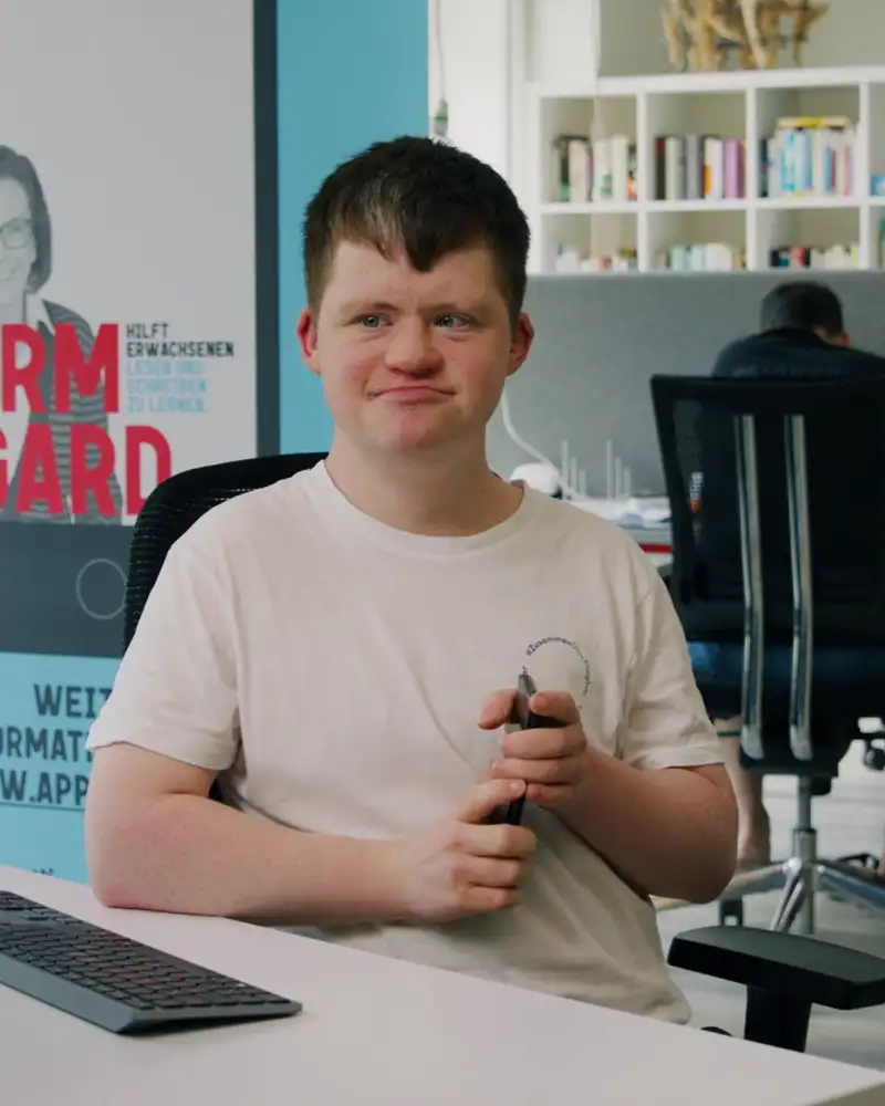 Ein junger Mann mit Behinderung sitzt in einem Büro am Schreibtisch. Er trägt ein weißes T-Shirt und schaut auf einen Computer-Monitor.