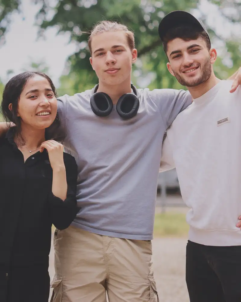 Zwei junge Männer und eine junge Frau stehen nebeneinander. Der Mann in der Mitte hat seine Arme um die beiden anderen gelegt.