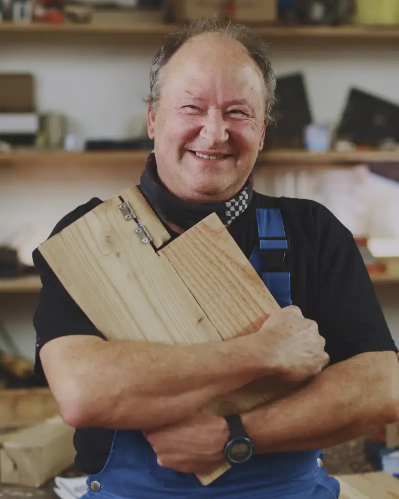 Ein Mann steht lächelnd in einer Werkstatt. Er hat Holzbretter in seinen Armen.