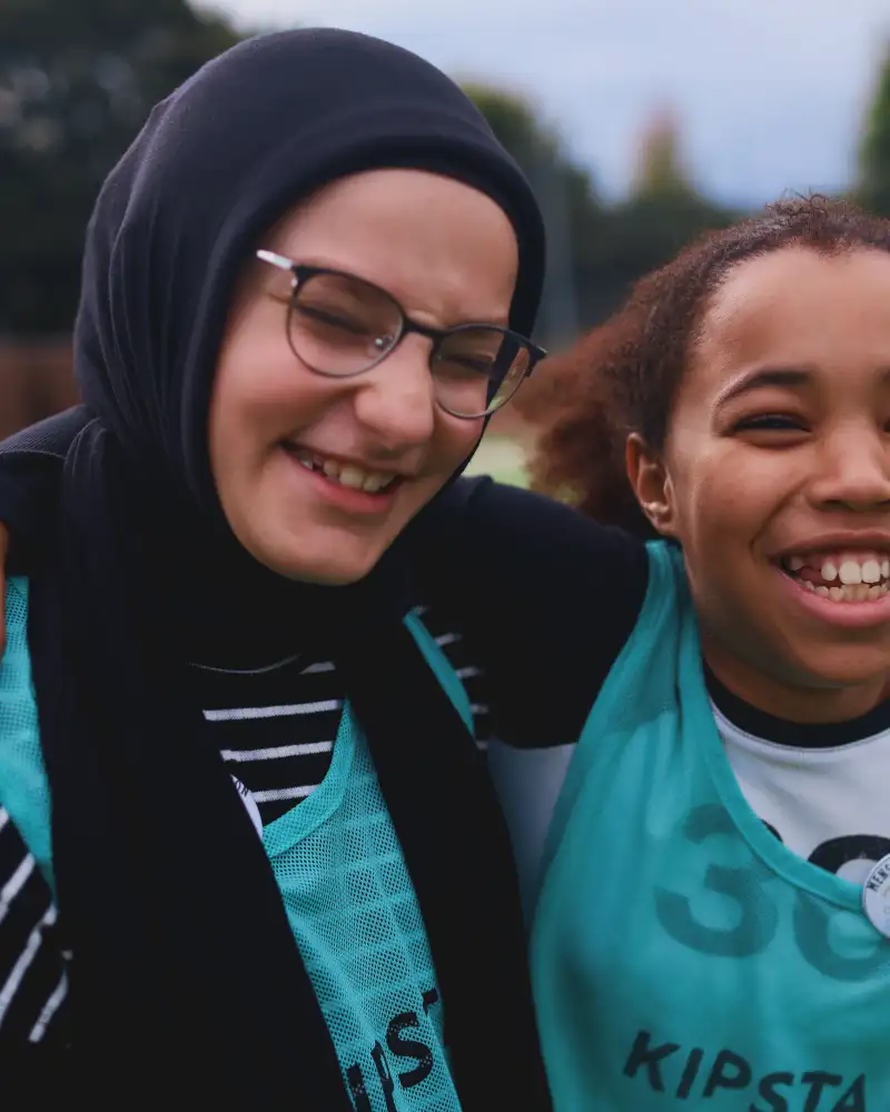 Zwei Mädchen stehen lachend auf einem Sportplatz und umarmen sich.