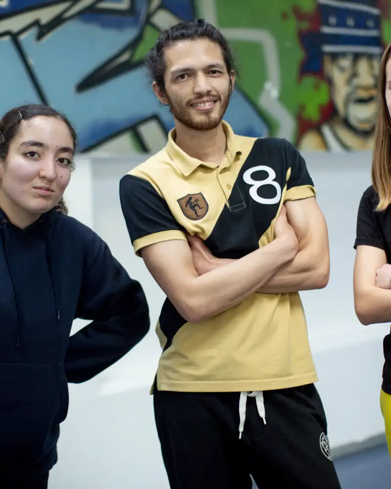 Ein junger Mann und zwei junge Frauen stehen in Sportkleidung in einer Sporthalle. Im Hintergrund ist eine mit Graffiti besprühte Wand.