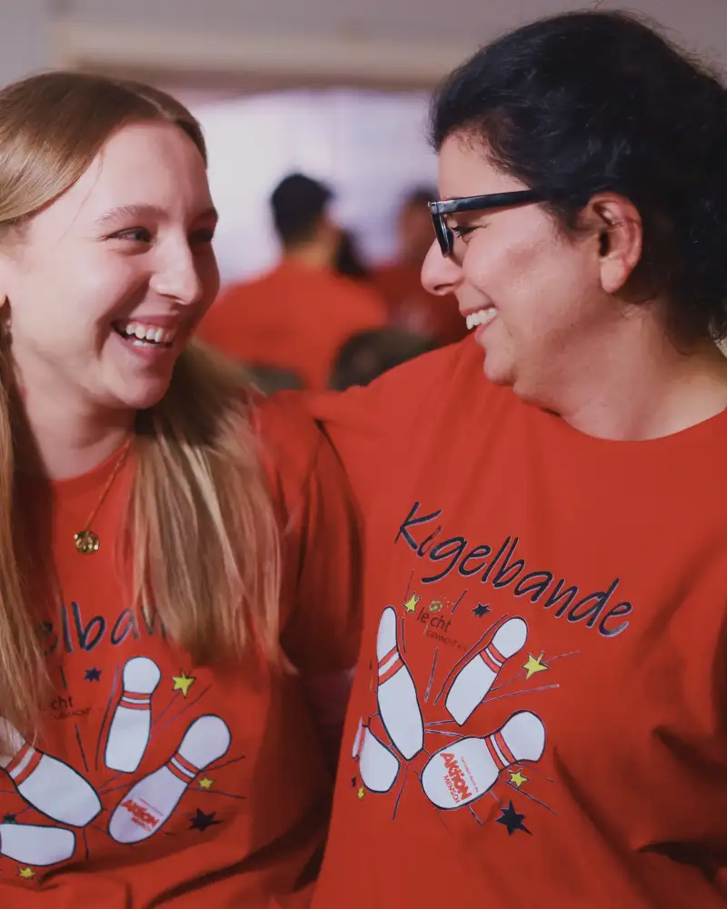 Zwei junge Frauen stehen Arm in Arm nebeneinander und lächeln sich an.