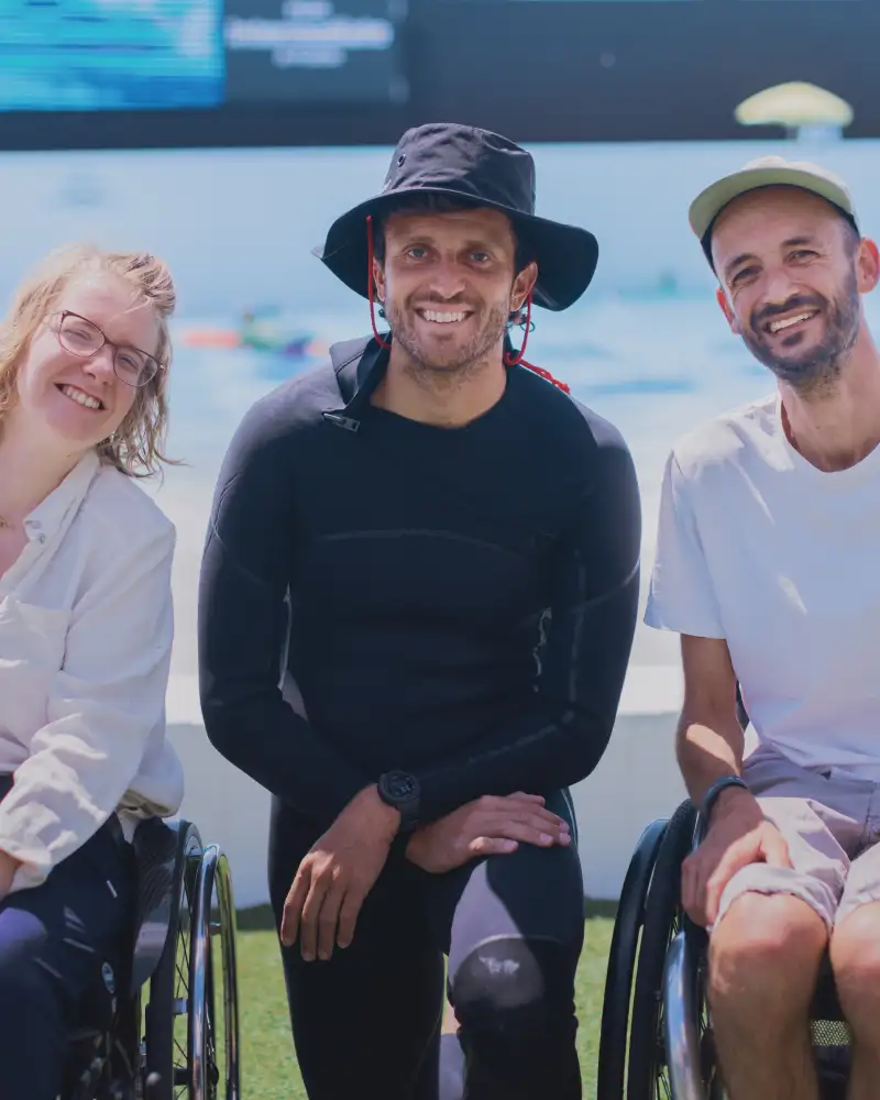 Eine junge Frau und zwei junge Männer lachend vor einem Schwimmbecken. Zwei von ihnen sitzen im Rollstuhl.