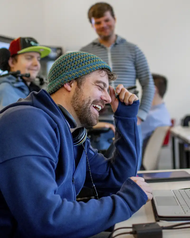 Ein Mann sitzt an einem Tisch vor einem Laptop und lacht herzlich. Um ihn herum sind drei weitere Personen, die zu ihm hinschauen.