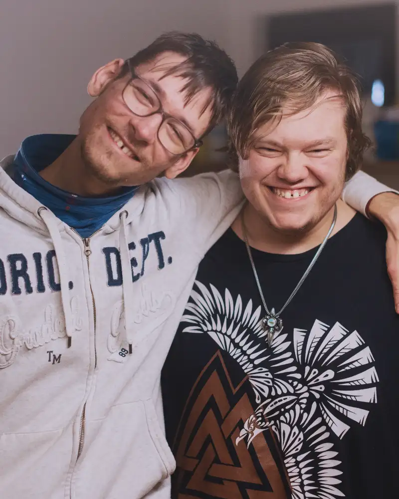 Zwei junge Männer stehen in einer Küche nebeneinander. Der eine hat seinen Arm um die Schultern des anderen gelegt. Beide lächeln.
