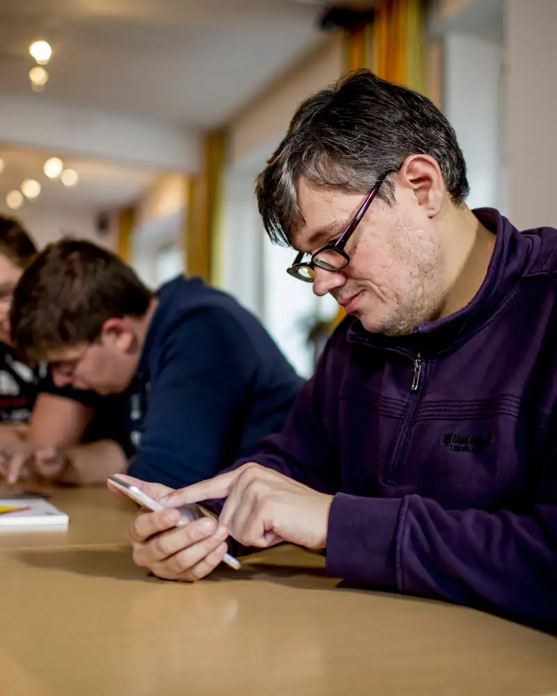 Ein Mann sitz an einem Tisch und schaut auf ein Smartphone. Neben ihm am Tisch sitzen zwei weitere Männer, die gemeinsam auf ein weiteres Smartphone gucken.