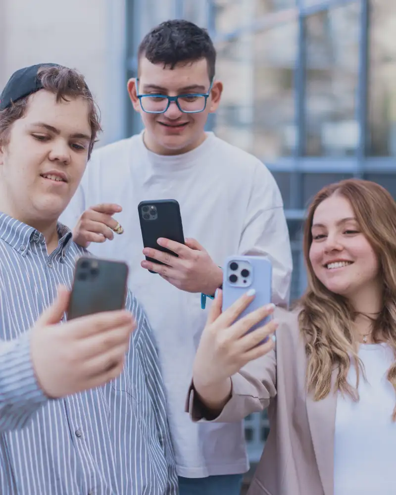Drei Jugendliche stehen nebeneinander. Alle halten jeweils ein Smartphone in der Hand und machen lächelnd Selfies von sich.