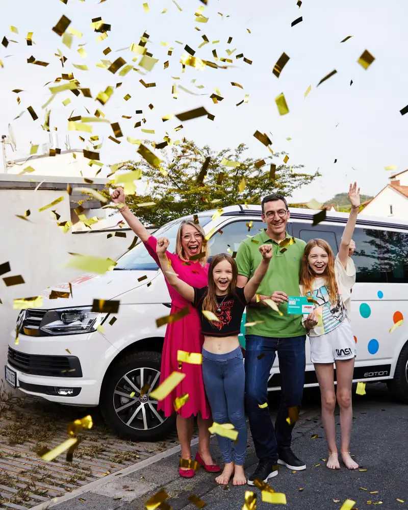 Eine Familie steht im goldenen Konfettiregen. Die Mutter und beiden Töchter recken jubelnd die Arme in die Luft, der Vater hält ein Glückslos in der Hand.