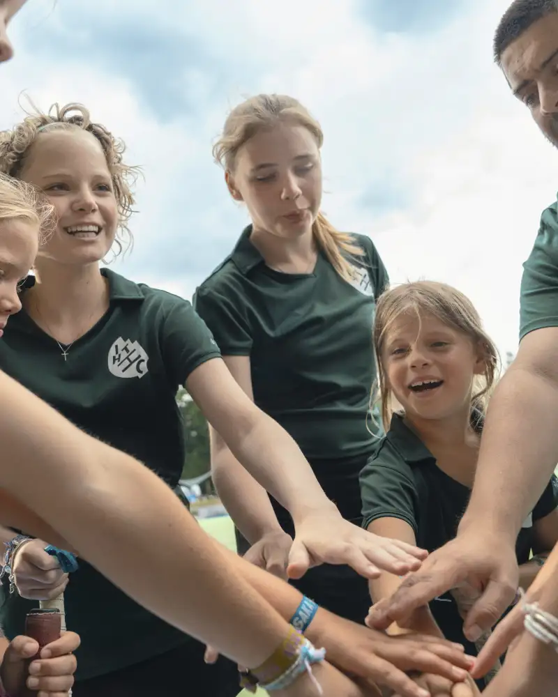 Eine Gruppe von Mädchen in grünen Sporttrikots mit ihrem Trainer. Sie schwören sich als Team ein, indem sie eine Runde bilden und alle ihre Hände aufeinandergelegt haben. Sie lächeln.