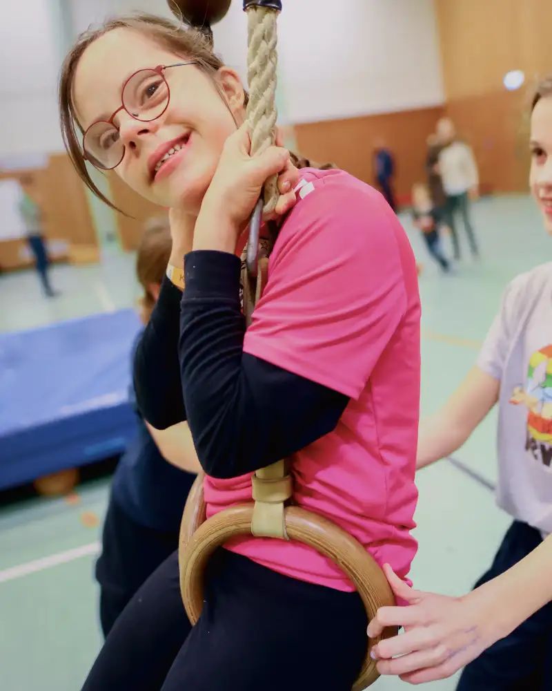 Zwei Mädchen in einer Turnhalle. Eine sitzt in Ringen, die andere schubst sie an.