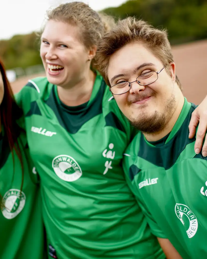 Drei Jugendliche stehen auf einem Sportplatz und umarmen sich. Sie alle tragen ein grünes Trikot.