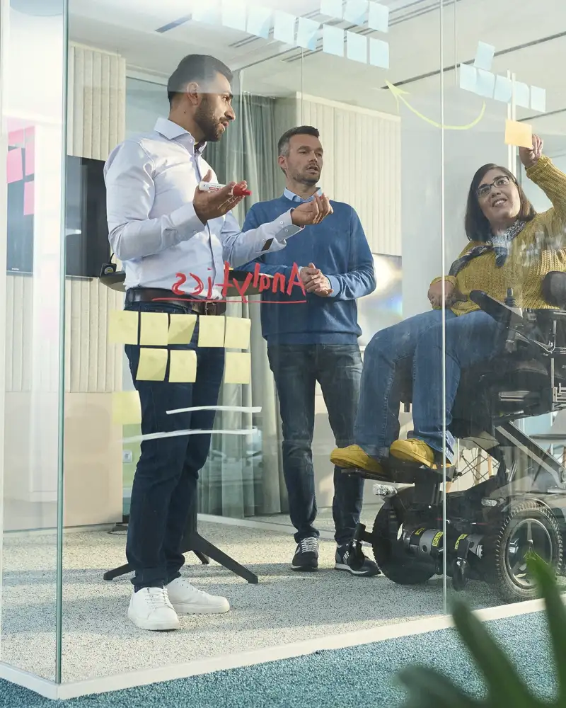 Drei Mitarbeitende der Aktion Mensch in einem Besprechungsraum. Der Besprechungsraum hat Glaswände, der Kamera-Blick ist von außen.