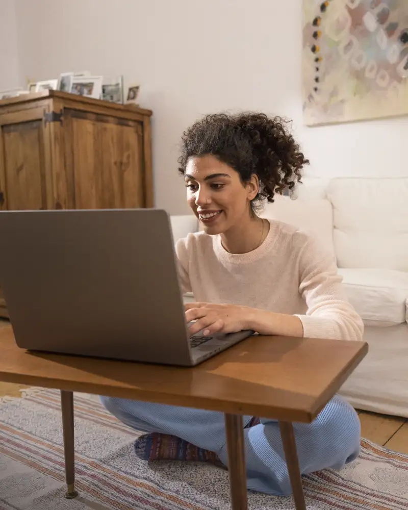 Eine junge Frau sitzt auf dem Boden ihres Wohnzimmers vor einem kleinen Tisch. Auf dem Tisch steht ein Laptop in den sie schaut.