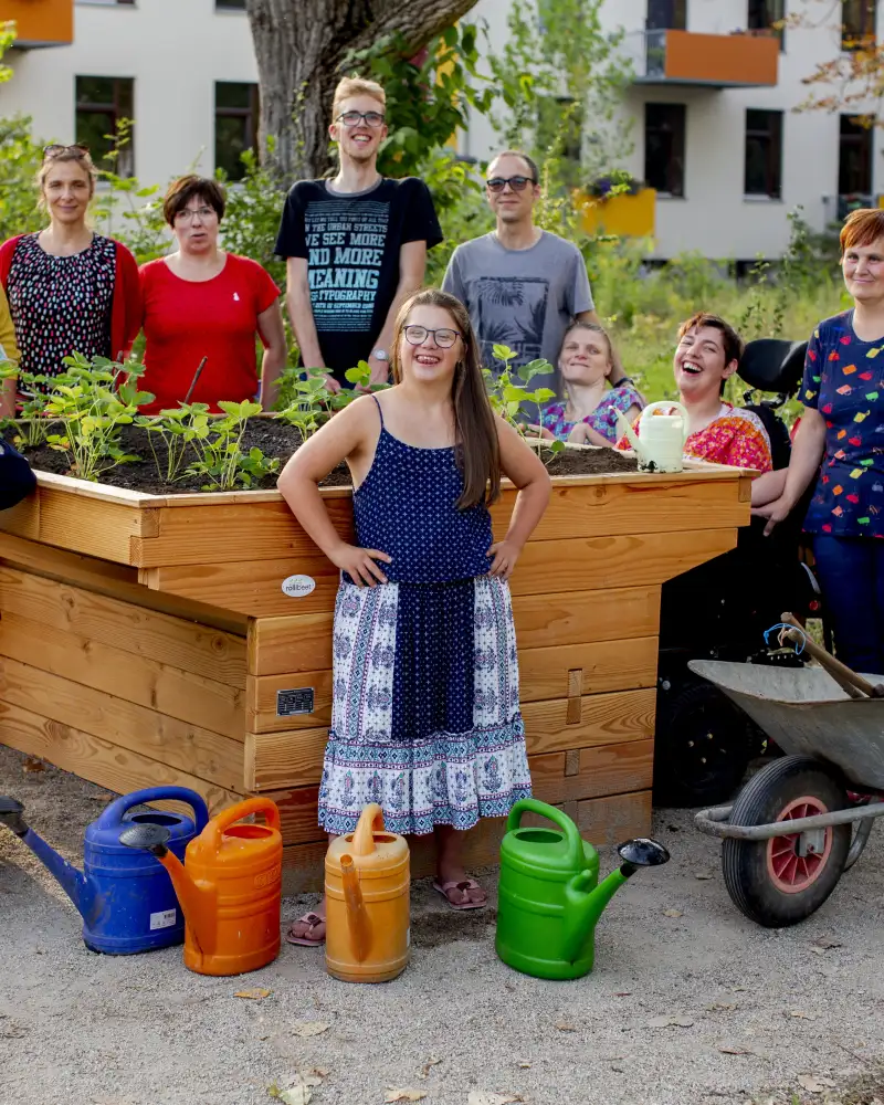 Eine Gruppe von Menschen mit und ohne Behinderung steht um ein Hochbeet, das sie gerade bepflanzt haben. Alle lächeln.