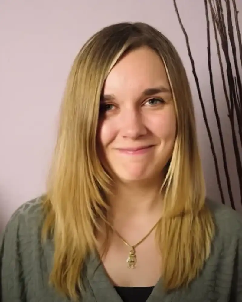 Eine junge Frau mit langen, blonden Haaren lächelt in die Kamera. Neben ihr steht ein Blumenstrauß.
