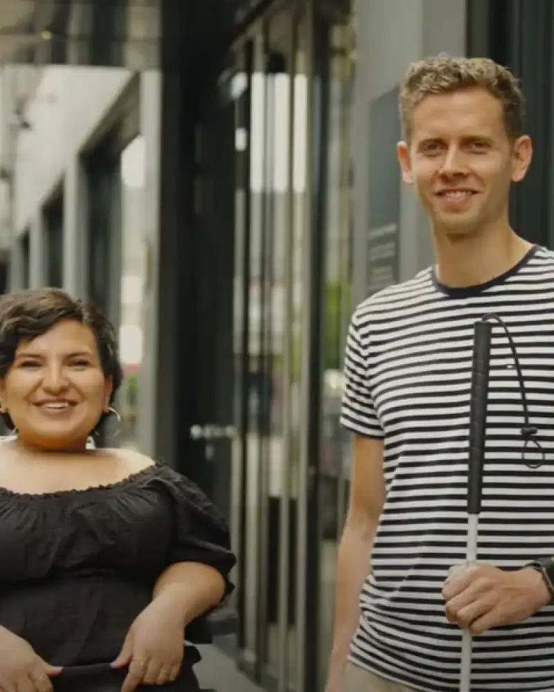 Eine junge Frau im Rollstuhl und ein junger Mann mit Blindenstock stehen in einer Fußgängerzone vor einem Eingang aus Glas.