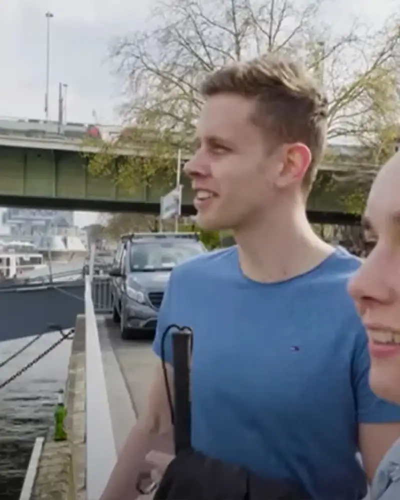 Ein junger Mann und eine junge Frau stehen am Rheinufer. Im Hintergrund sieht man eine Straßenbahn über eine Brücke fahren.