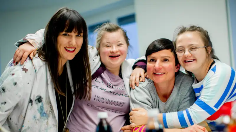 Vier junge Frauen stehen nebeneinander, umarmen sich und lachen in die Kamera.