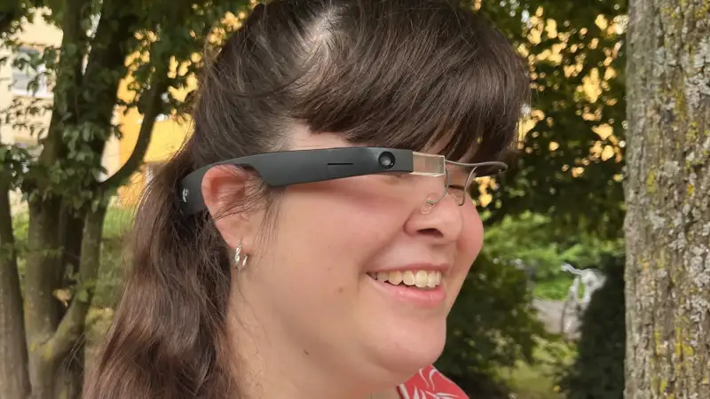 Junge Frau mit langen dunklen Haaren und sommerlicher roter Bluse trägt ein Brillengestell, in dessen rechten Bügel eine Kamera, ein kleines Display und weitere Technik integriert sind.