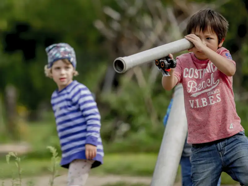 Zwei Kinder spielen. Ein Junge benutzt ein schmales Rohr wie ein Fernrohr. So als suche er etwas.