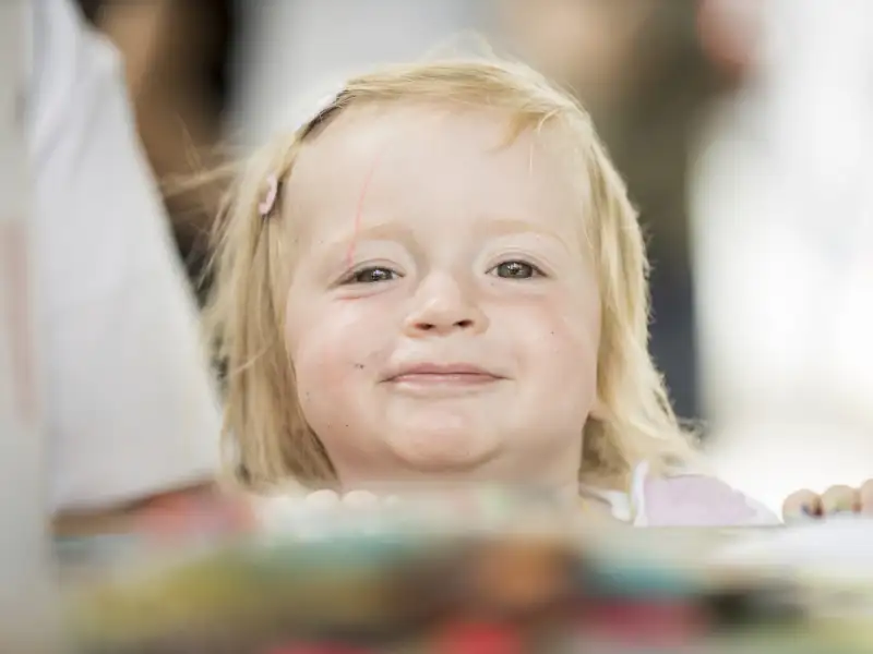 Ein kleines blondes Mädchen mit Spange im Haar schaut neugierig über einen Tisch hinweg direkt in die Kamera.