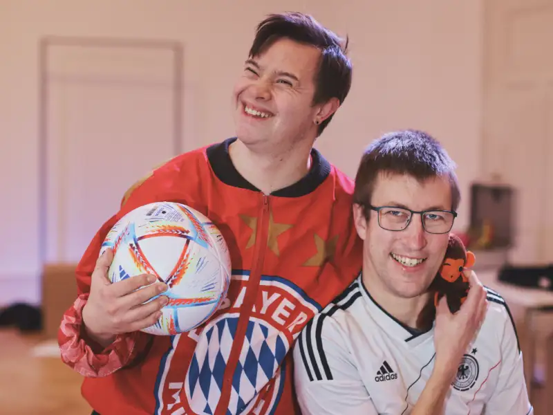 Zwei junge Männer in Sporttrikots stehen lachend nebeneinander. Der eine hält eine Fußball unter dem Arm.