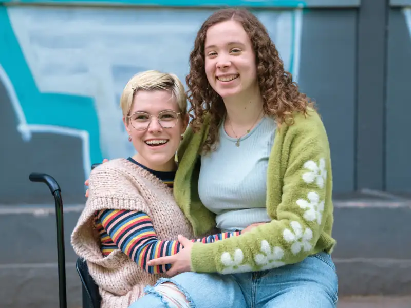 Zwei junge Frauen umarmen sich freundschaftlich. Eine von ihnen sitzt im Rollstuhl.