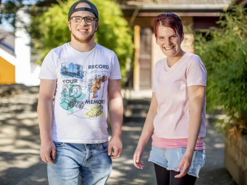 Okan und Zora. Zwei junge Menschen lachen in die Kamera. Okan hat einen rötlichen Bart und eine Brille. Er trägt ein nach hinten verdrehtes Basecap. Zora ist eine junge Frau mit kurzen, dunklen Haaren.