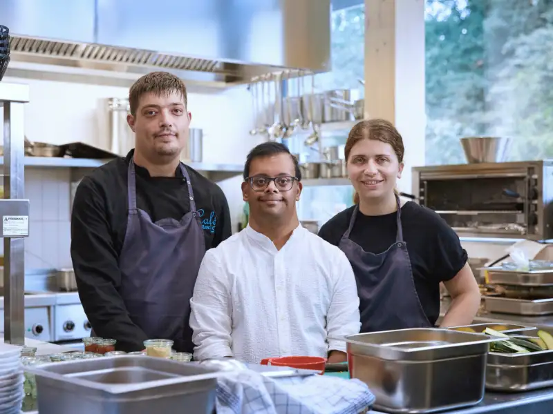Drei junge Erwachsene unterschiedlicher Herkunft stehen in einer Café-Küche.