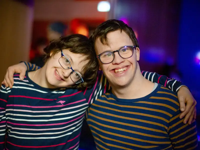 Zwei junge Menschen liegen sich in den Armen und lächeln in die Kamera.