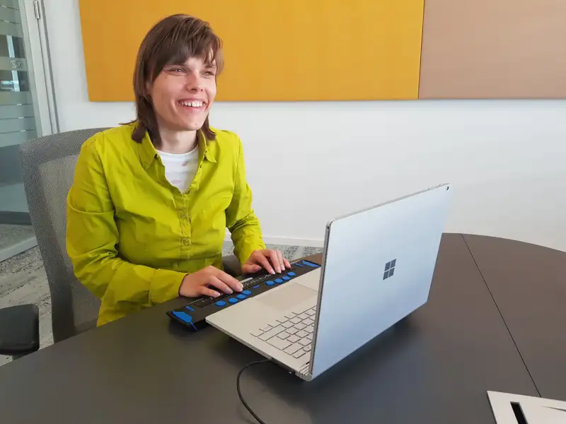 Eine blinde junge Frau sitzt lächelnd vor einem Laptop mit Braillezeile.